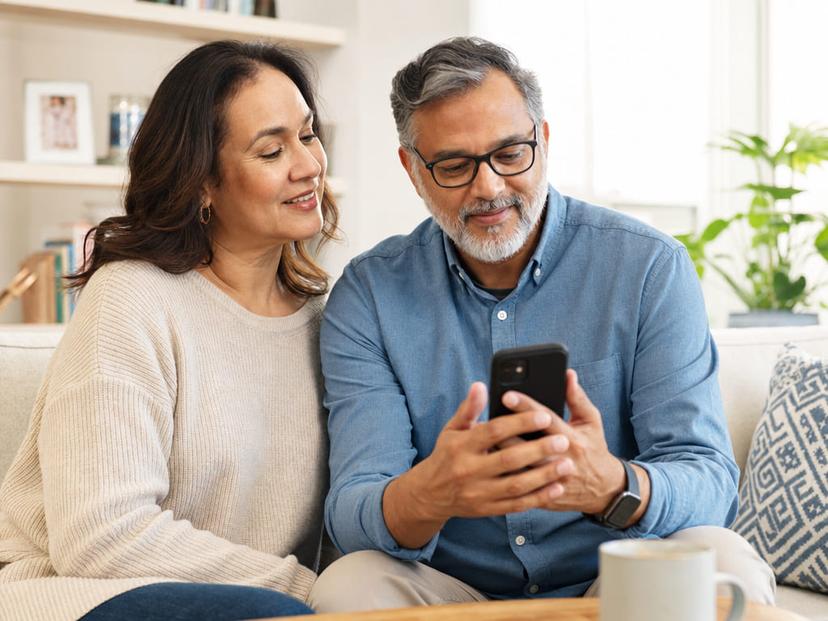 A couple sitting together on a sofa at home, looking at a health app on a smartphone